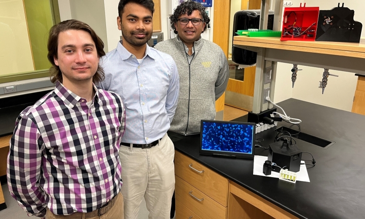 Three men stand in a lab next to equipment on a black table top.