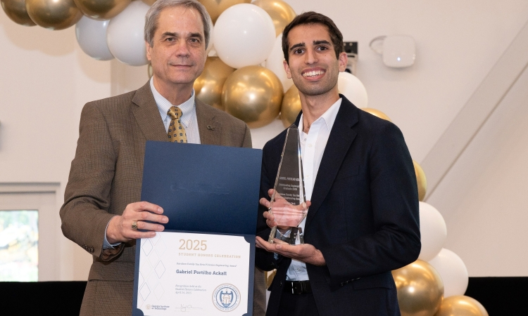 Two men stand in front of a gold and white balloon arch holding a trophy and certificate