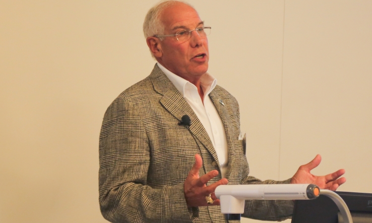 Ron Blum in a plaid jacket and a white dress shirt speaking at a lectern and gesturing with his hands.