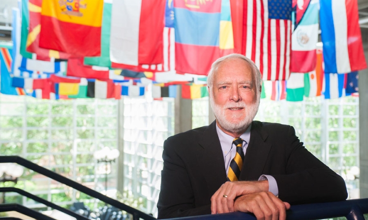 Georgia Tech President Emeritus G. Wayne Clough stands with his arms resting on a stair handrail with international flags hanging in the background.