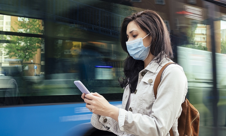 A woman wearing a blue surgical map looks at her smartphone as a bus drives by
