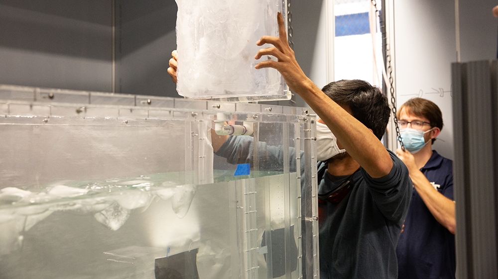 A man lowers a block of ice into a tank of water as another man in a surgical mask looks on