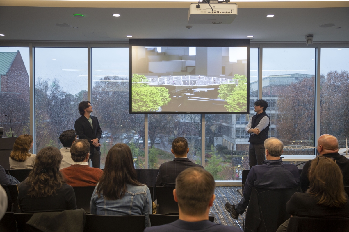 two men look at a projection screen in front of a seated crowd