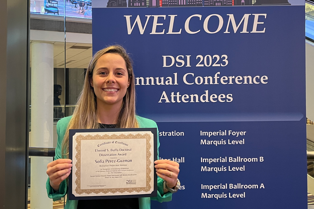 A woman holds a certificate in front of a conference welcome sign