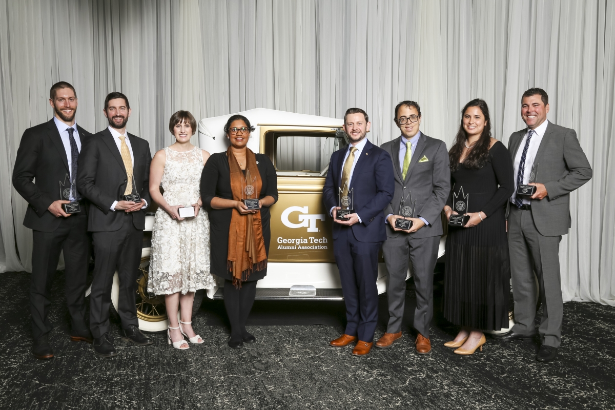 A group of people poses in front of an antique gold car and a white curtain 