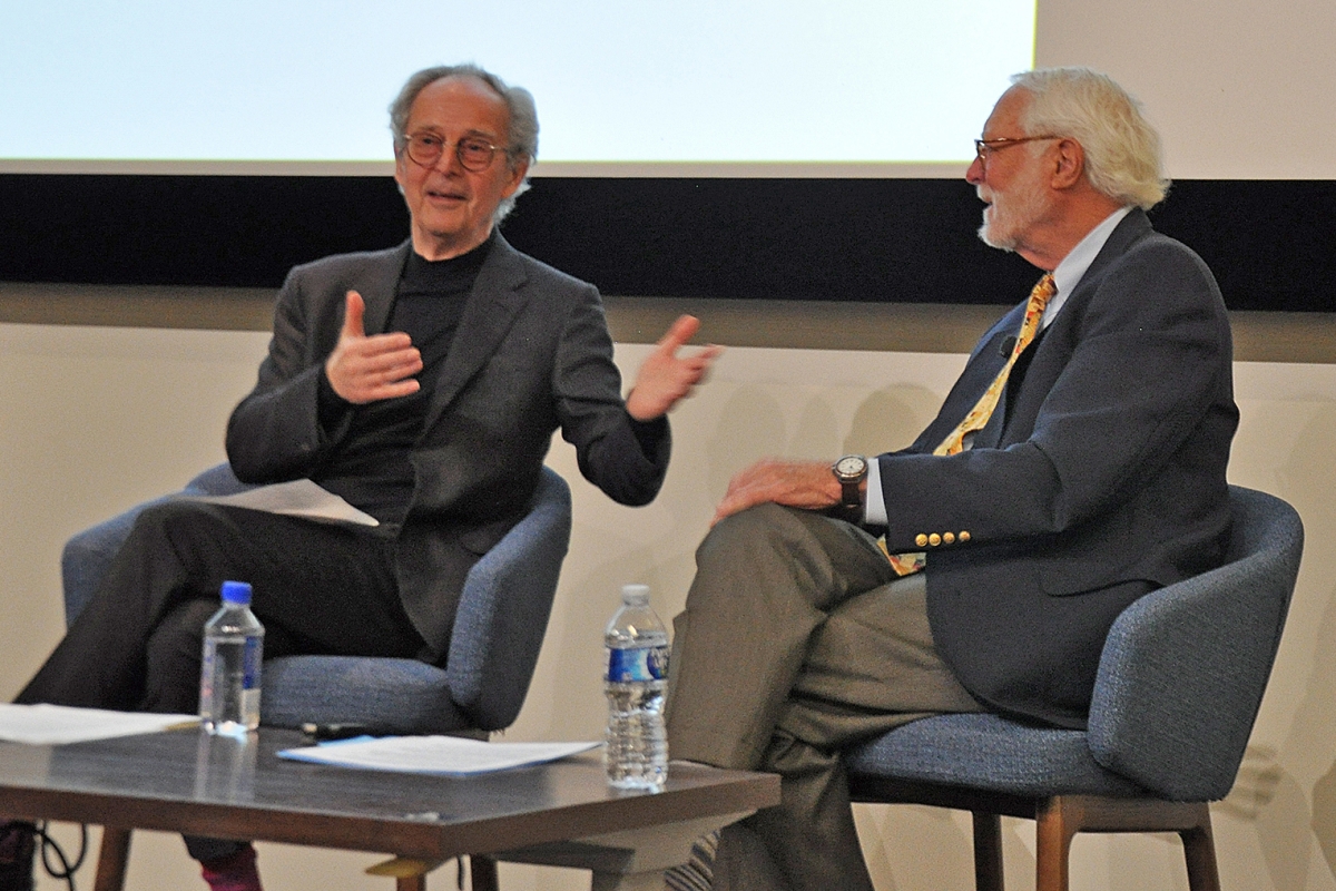 Two men in conversation sit side by side on blue chairs 