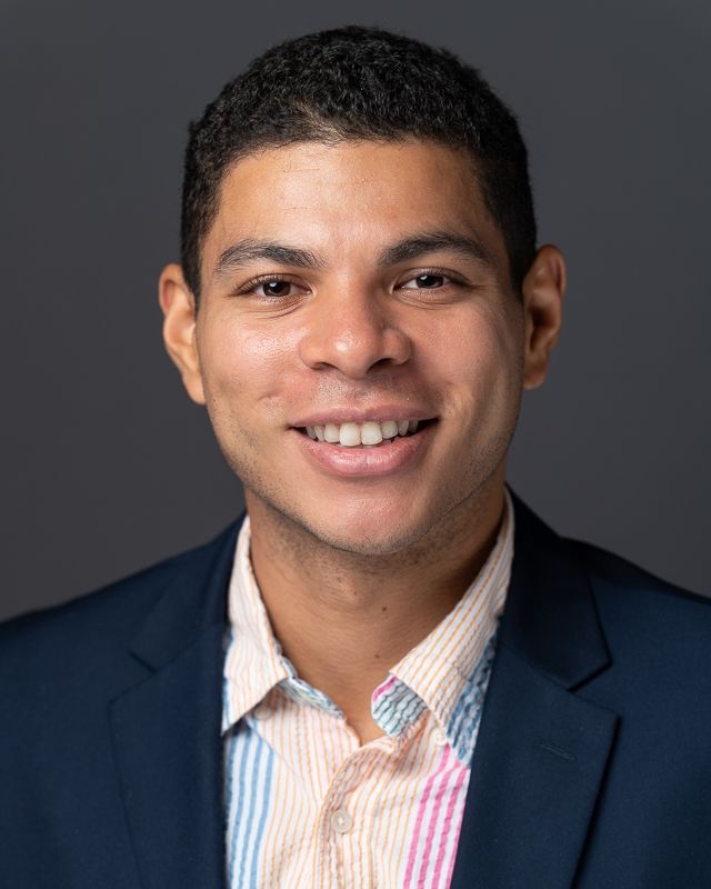 A portrait of PhD student Christian Douglas in a blue blazer and multi-colored striped oxford shirt