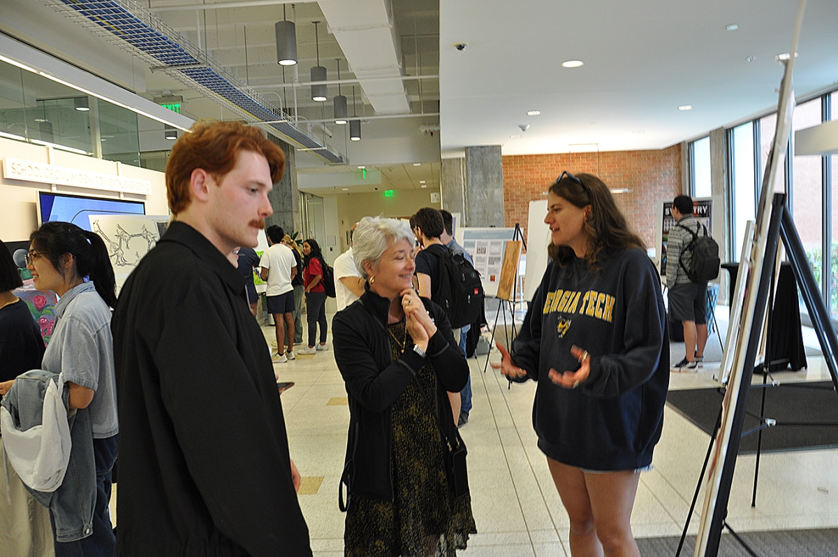 Woman gazes at project on easel which is being explained by a red haired male student on her left and a woman student on her right