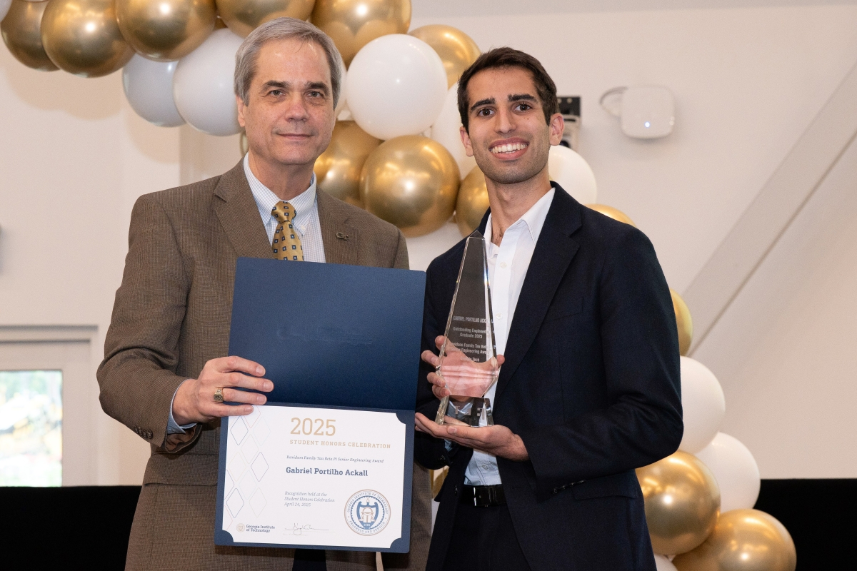 Two men stand in front of a gold and white balloon arch holding a trophy and a certificate.
