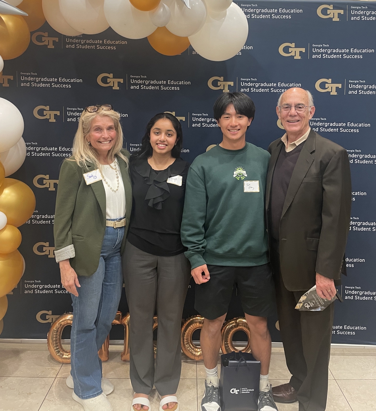 Professor Sandi Bramblett, students Deeya Mepani and Joe Zheng and Rafael Bras pose in front of a blue Georgia Tech background and white and gold balloons.