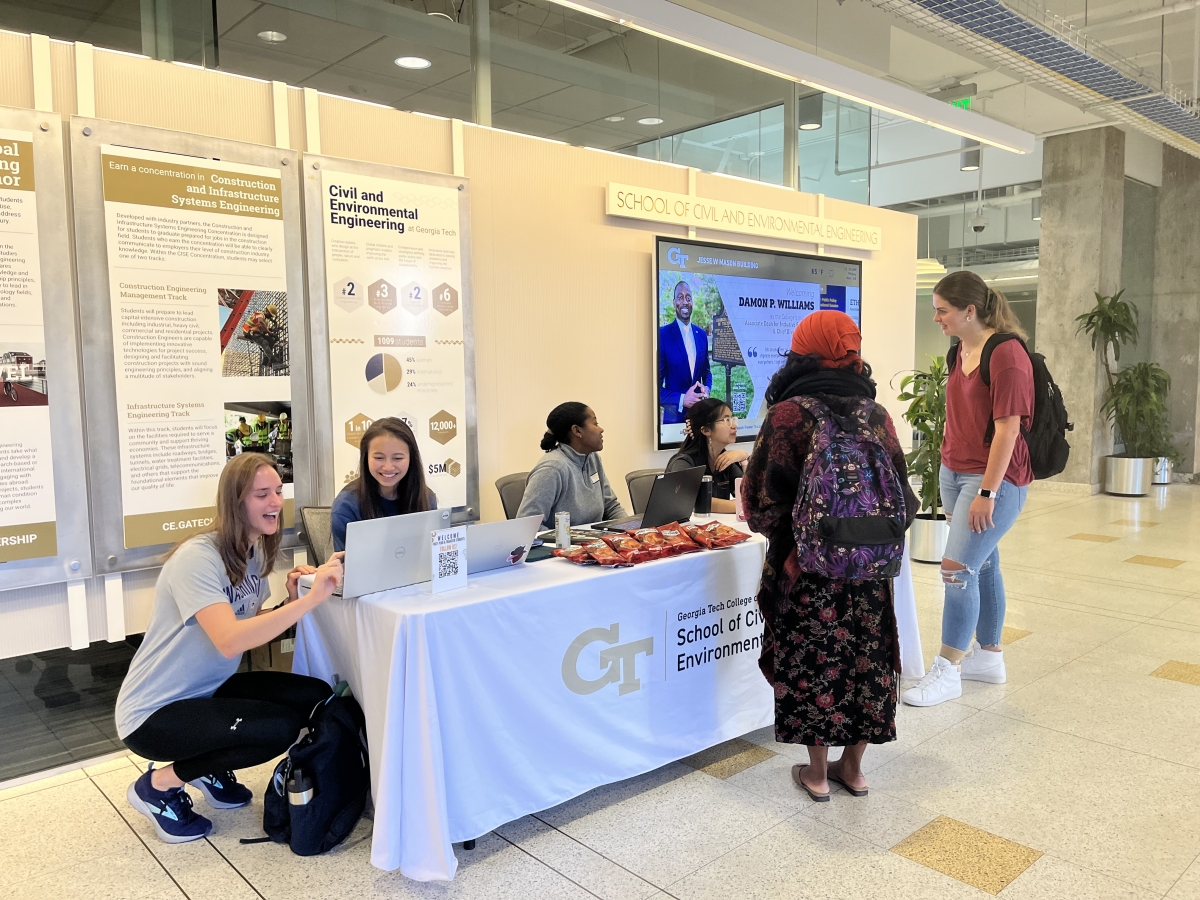 A group of student Ambassadors sitting behind a table with a white tablecloth in Mason lobby engage with two female students