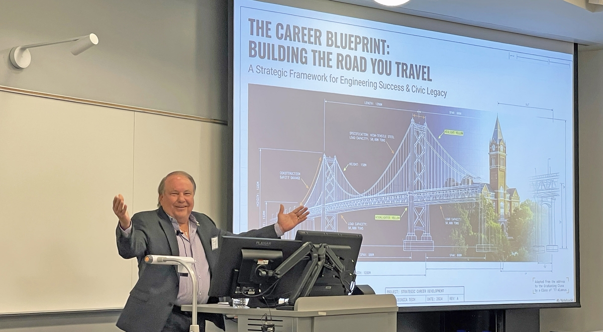 John Carroll with his arms raised in a navy blue blazer and lavender shirt speaking to students