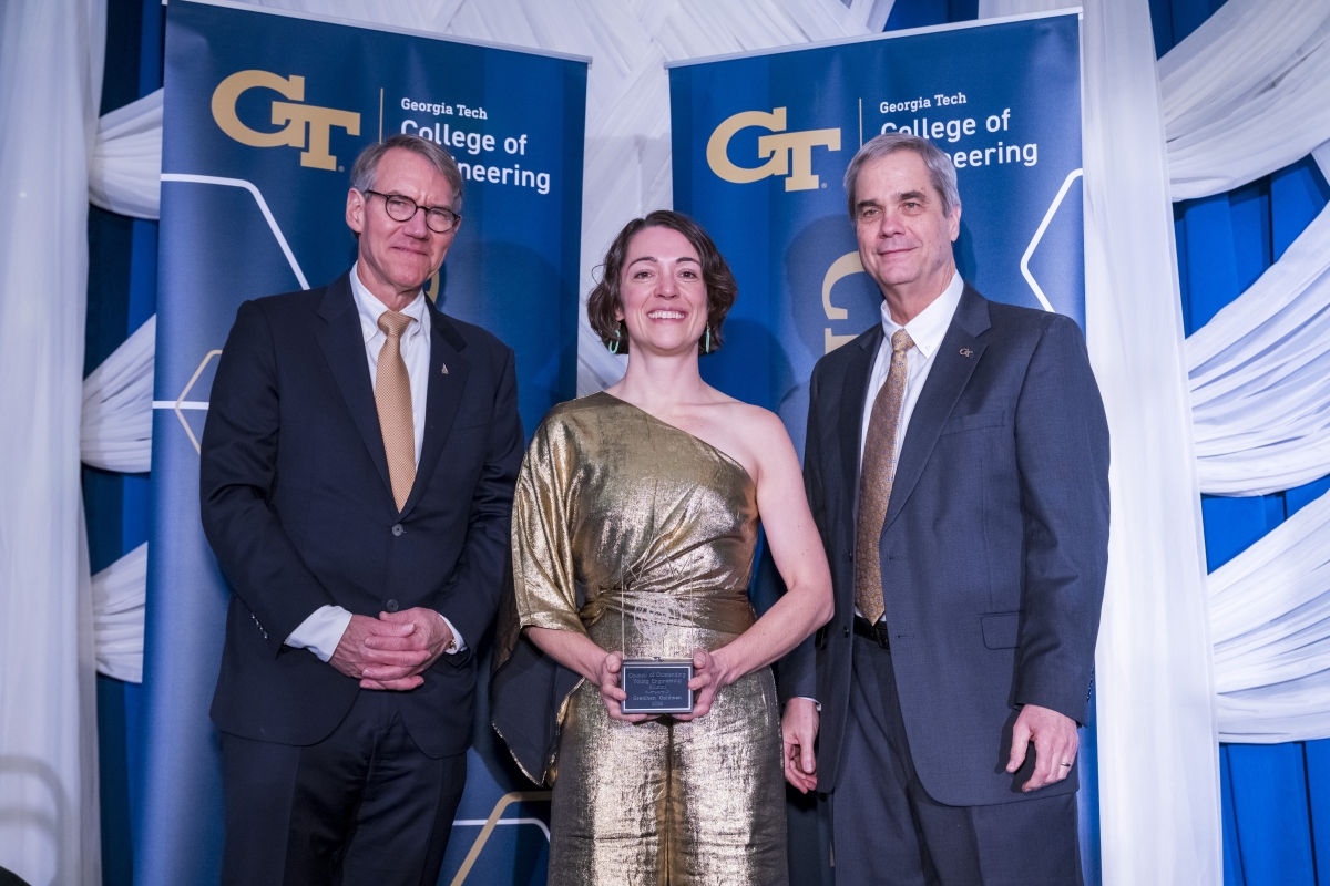 A woman in a gold dress holds an award between two men in suits