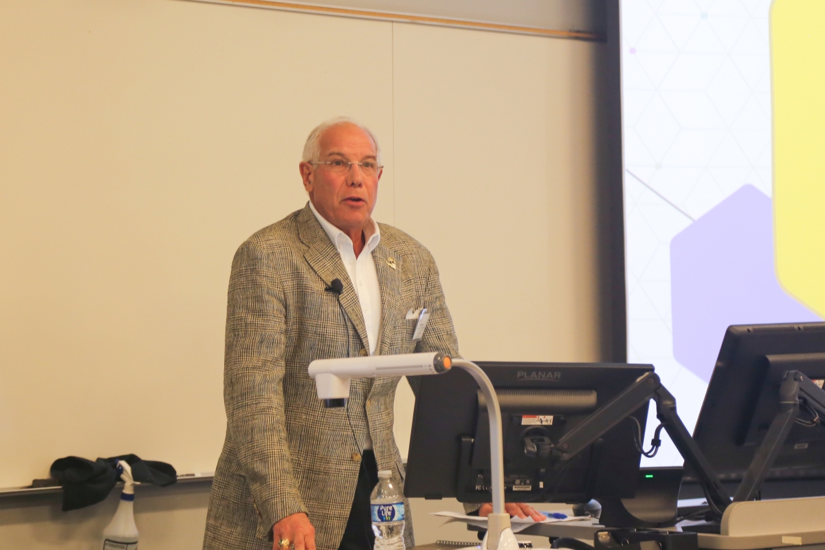 Ron Blum in a brown plaid jacket and white dress shirt speaking at a lectern with both of his hands placed on the lectern