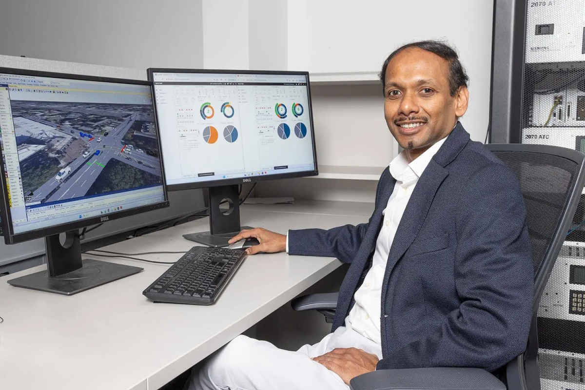 A man sits next to two computer monitors showing pie graphs and a road intersection