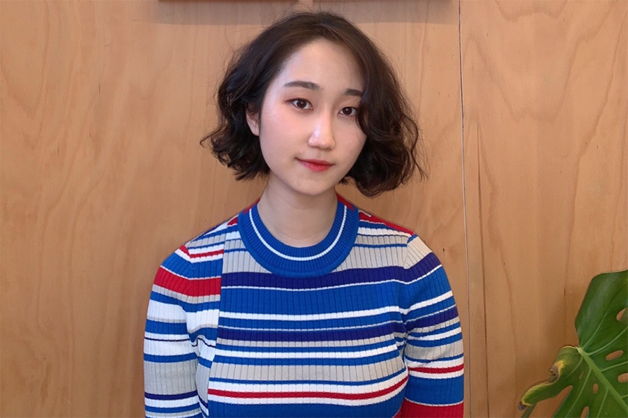 Laura Yang wears a striped sweater and poses in front of a wooden background with a plant visible in the lower right corner