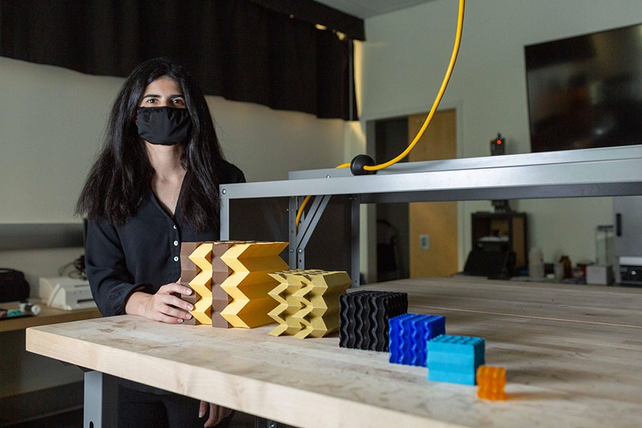 Larissa Novelino, a Ph.D. student at Georgia Tech, has long black hair and is wearing a black shirt and a black mask ovrer her nose and mouth. She is standing next to a table showing several metamaterial prototypes at different scales. They are different colors and range in size from smallest to largest.