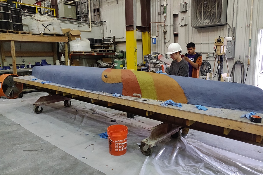 A person in a white hard hat works on a blue canoe with red orange and yellow stripes 