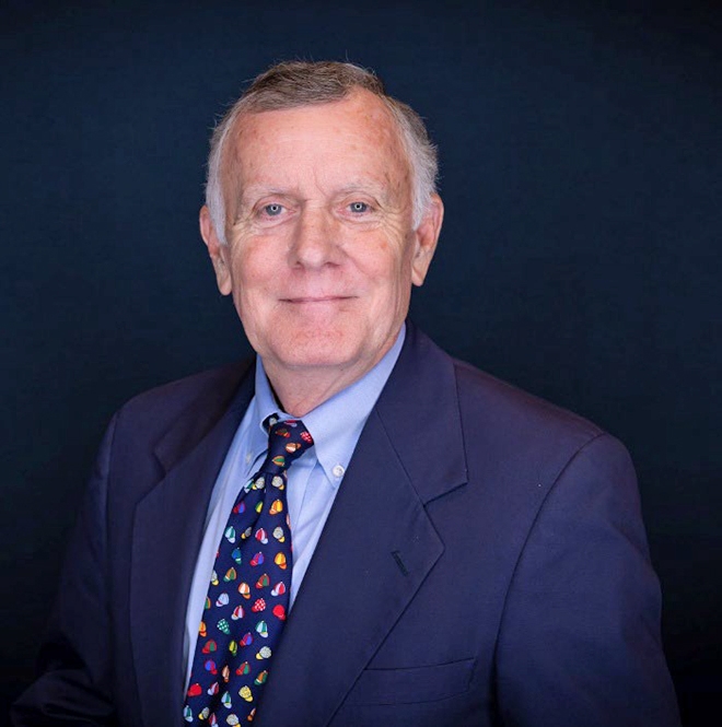 Portrait of Bill Daniel in a navy blue suit with a light blue suit and a blue tie with multi-colored objects on it